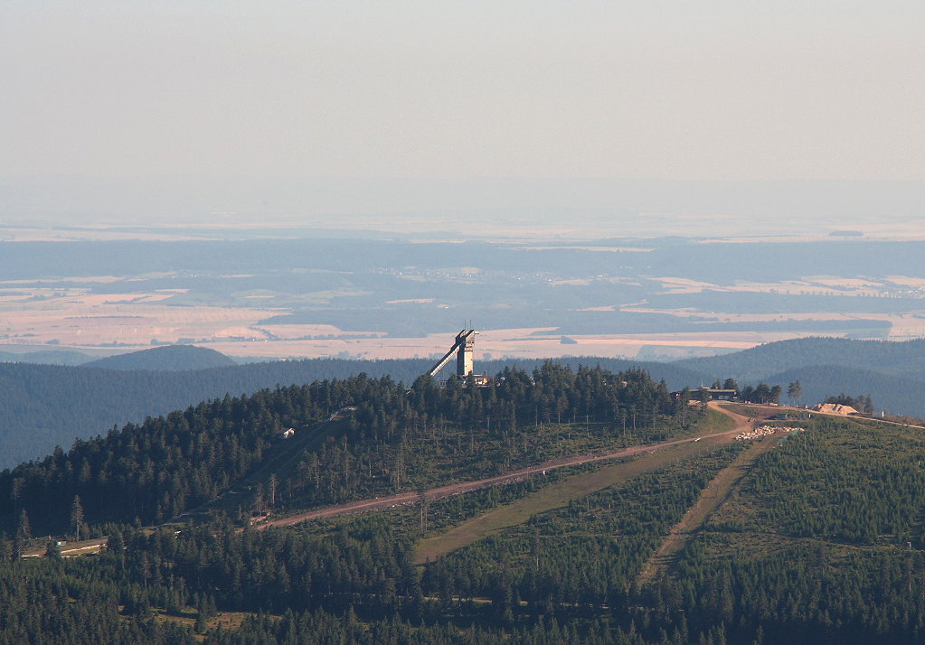 Der Gipfel des Wurmbergs mit dem ca. 30 m hohen Turm der Skisprungschanze, dahinter ber den letzten Gipfeln des Sdharzes die Hainleite in Thringen mit ihrem Wechsel von Feldern und Wald, darber bzw. dahinter zart am Horizont die hchsten Berge des Thringer Walds, ca. 120 - 130 km entfernt; Blick am frhen Abend des 21.07.2013 vom Gipfelrundweg des Brocken Richtung Sden...