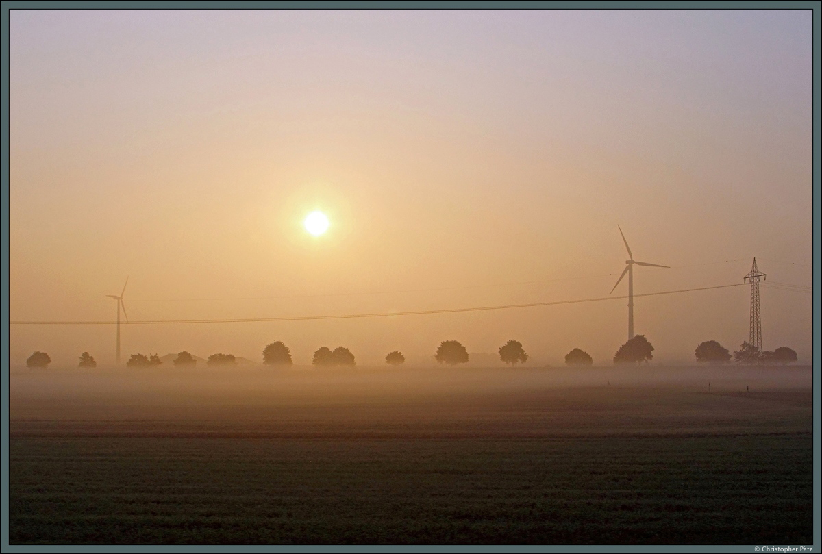 Langsam löst sich am 21.08.2015 der Morgennebel auf, als sich die aufgehende Sonne über die Felder und Windräder bei Eilvese erhebt. 