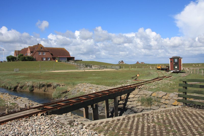 Hallig Nordstrandischmoor - 16.06.2008 - Fast wie in einer Fluchtlinie sind die Warften angeordnet, hier die Neuwarft im Vordergrund und davor abgestellte Fahrzeuge der Halligbahn.   