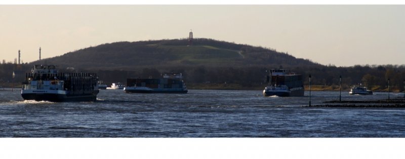 Rush Hour auf dem Rhein - Fotografiert vom Rheinufer Duisburg-Bruckhausen Blickrichtung flussaufwrts. Kann mir jemand sagen, was das fr ein Hgel im Hintergrund ist, auf dem eine Skulptur (Mann) steht?